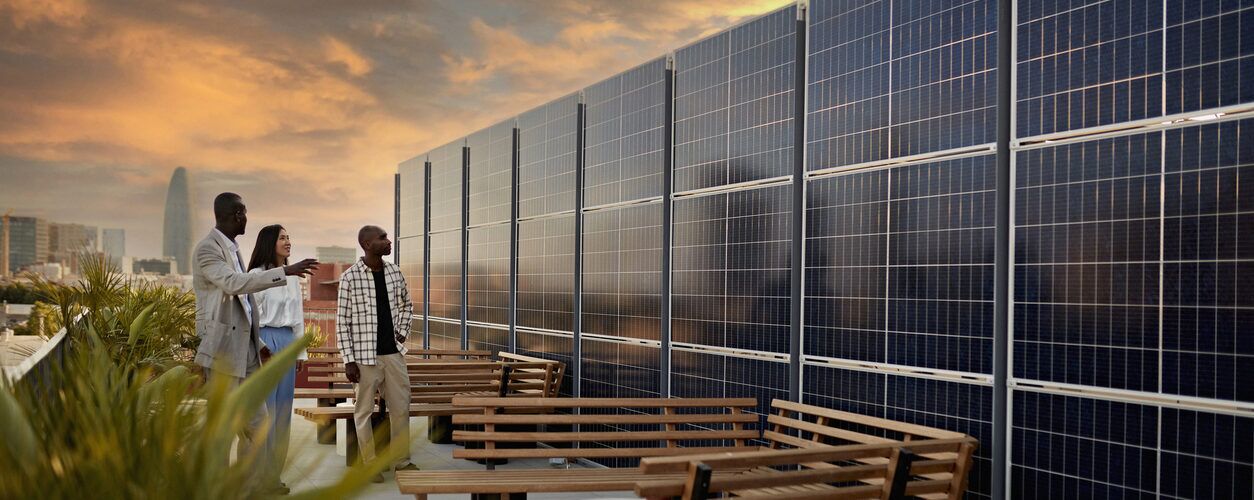 Three people standing on a rooftop with benches, looking at large vertical solar panel installations at sunset with a city skyline in the background.